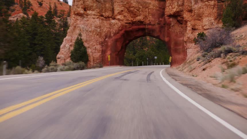 A point-of-view driving shot emerges from a tunnel and drives along a winding road towards another red rock tunnel in the desert