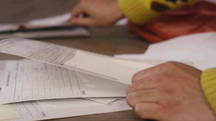 A person's hands slowly sort through a stack of official-looking documents on a wooden desk