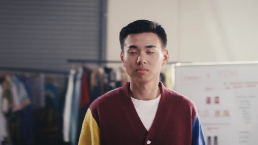 Portrait of a serious young Asian fashion designer looking at the camera in his creative studio with clothing racks in the background