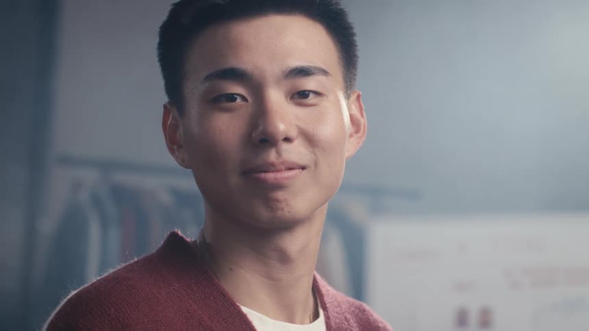 Close-up portrait of a young, confident Asian man smiling kindly at the camera in a hazy, softly lit room