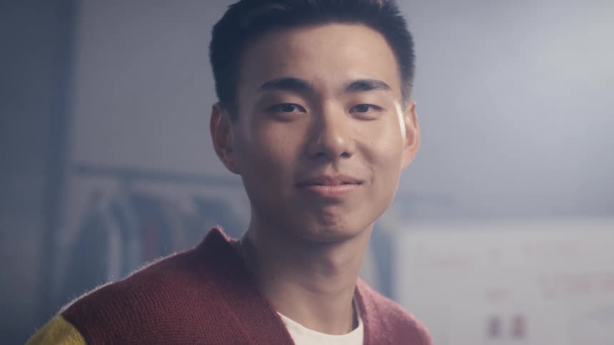 A handsome young Asian man in a colorful sweater looks directly at the camera with a confident smile in a hazy, backlit room