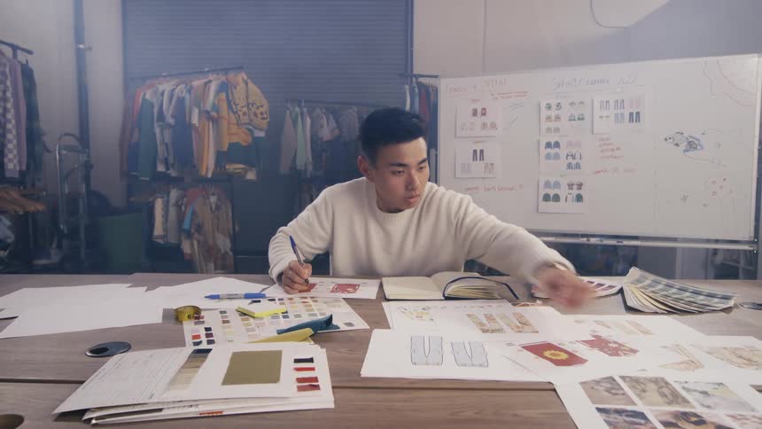 A focused young Asian fashion designer sketches new designs and organizes his mood board at a large table in his creative studio