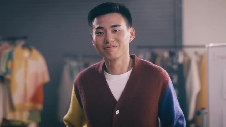 A confident young Asian man buttons his trendy, multi-colored cardigan and smiles warmly at the camera in a clothing studio