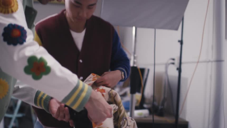 Two male stylists meticulously prepare a red floral-patched varsity jacket for a photoshoot in a professional studio setting