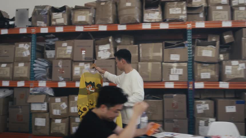 A young fashion designer inspects colorful sweaters on a clothing rack in a busy warehouse stockroom