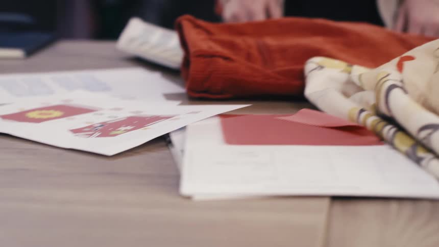 A fashion designer's hands move across a creative desk, working with color swatches, sketches, and fabric samples in a studio