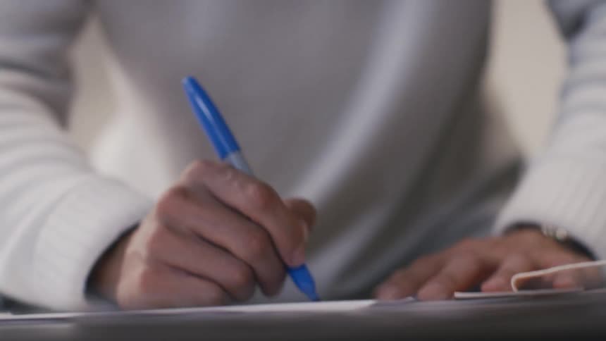 A young man in a white sweater writes on paper with a blue marker while planning a creative project in slow motion