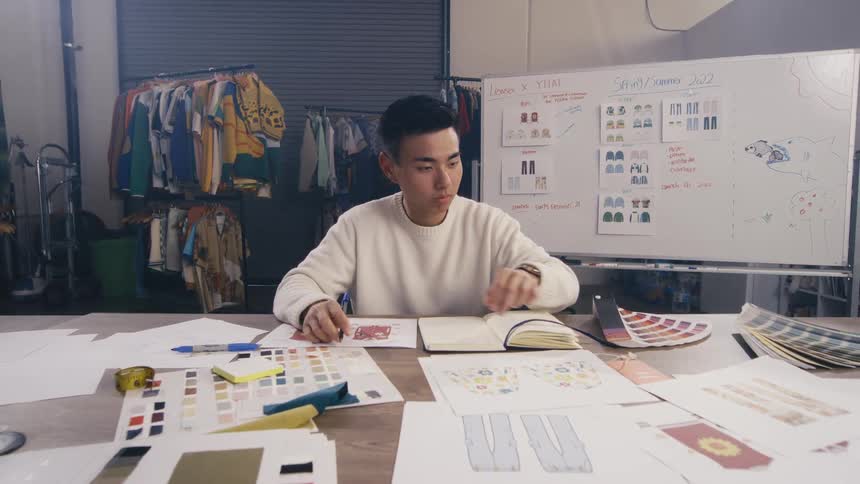 A young Asian fashion designer meticulously works on new clothing designs, surrounded by sketches and color swatches in his creative studio
