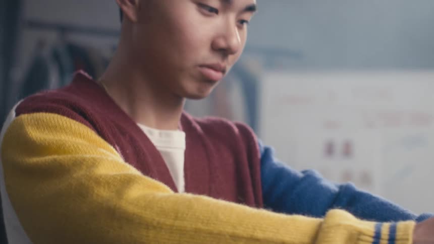 A slow tilt-up shot reveals a stylish young Asian man in a colorful varsity cardigan who looks confidently at the camera