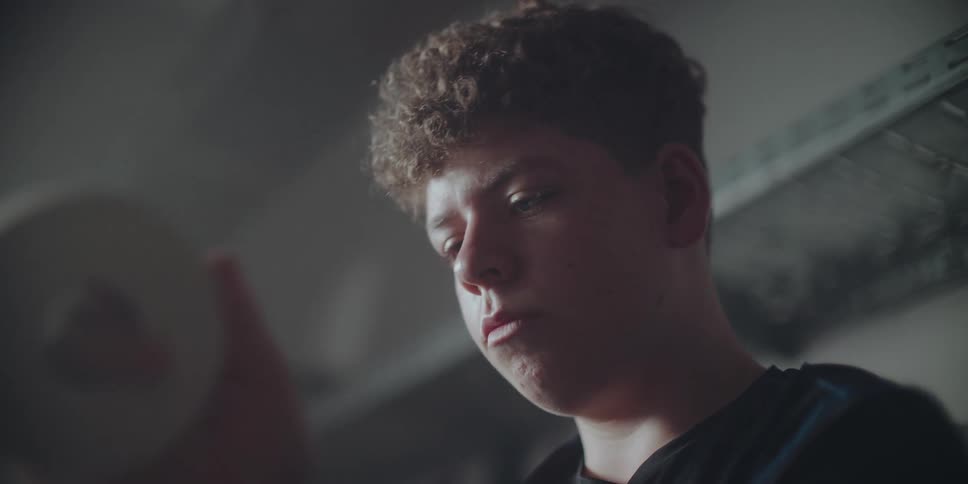 A determined young man with curly hair focuses intently on his work in a moody, dimly lit workshop