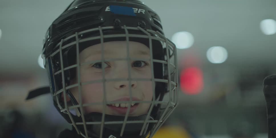 A smiling young boy in a full hockey helmet looks directly at the camera in a close-up shot