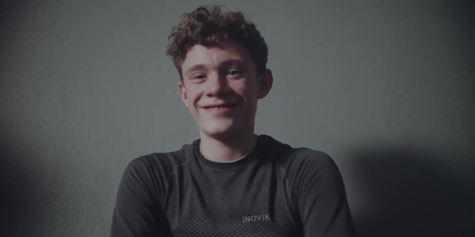A happy young man with curly brown hair in an athletic shirt smiles for a portrait against a plain wall
