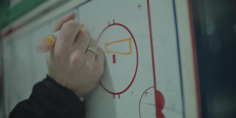 A hockey coach draws out a play on a white board with an orange marker in slow motion