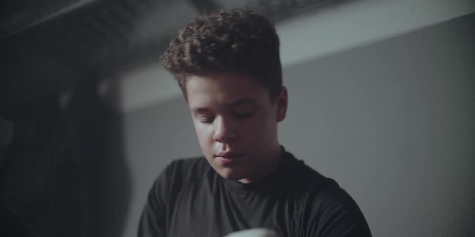 A focused teenage boy with curly hair carefully pours from a white container in a dimly lit garage setting