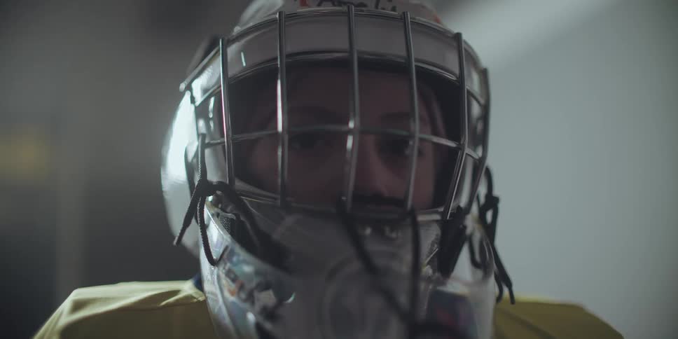 An intense close-up of a determined young hockey goalie staring directly into the camera from behind a face mask