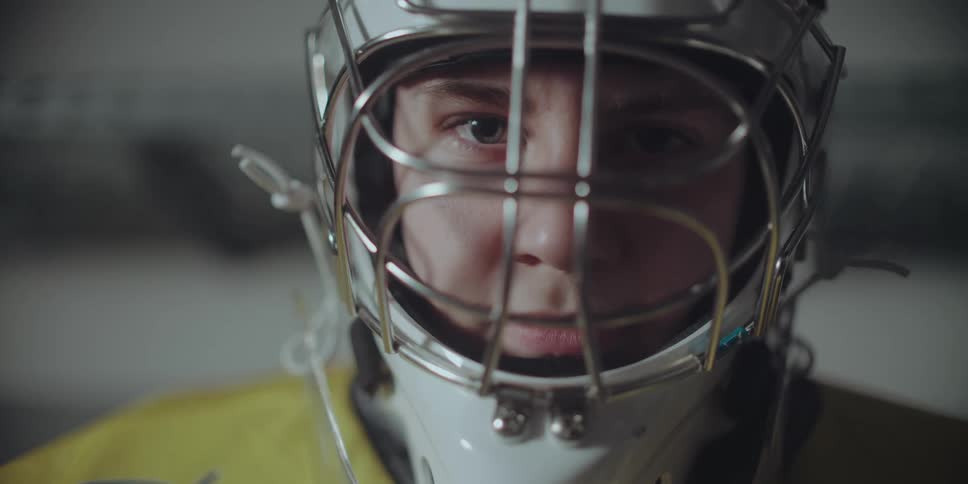 An intense close-up of a focused young hockey goalie staring directly into the camera through their protective helmet