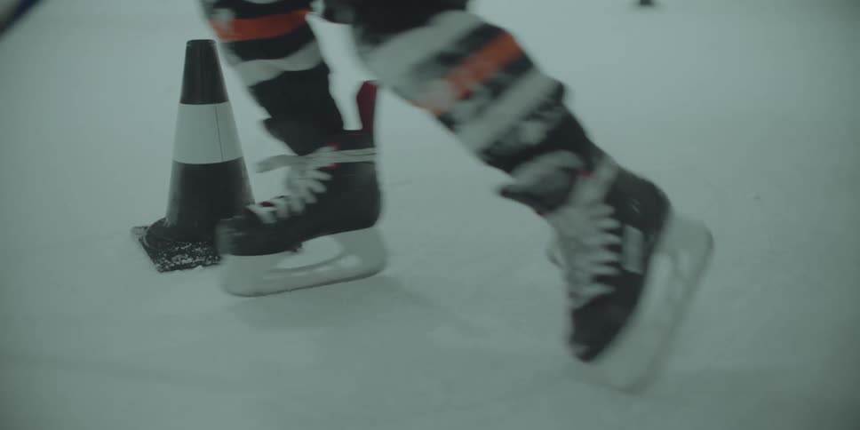 A close-up slow-motion shot of a hockey player's skates and stick maneuvering a puck around a cone on the ice