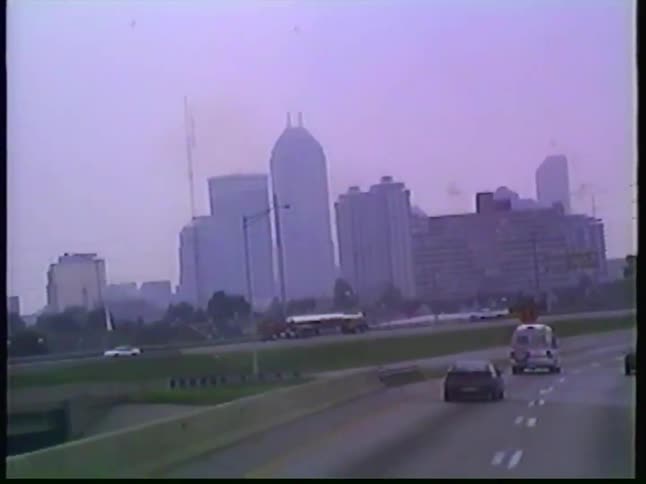 A vintage point-of-view shot from a vehicle driving on a highway toward the hazy Indianapolis city skyline