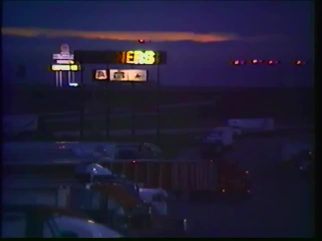 A grainy, vintage pan across a busy truck stop at twilight, with the glow of neon signs and highway lights dotting the landscape