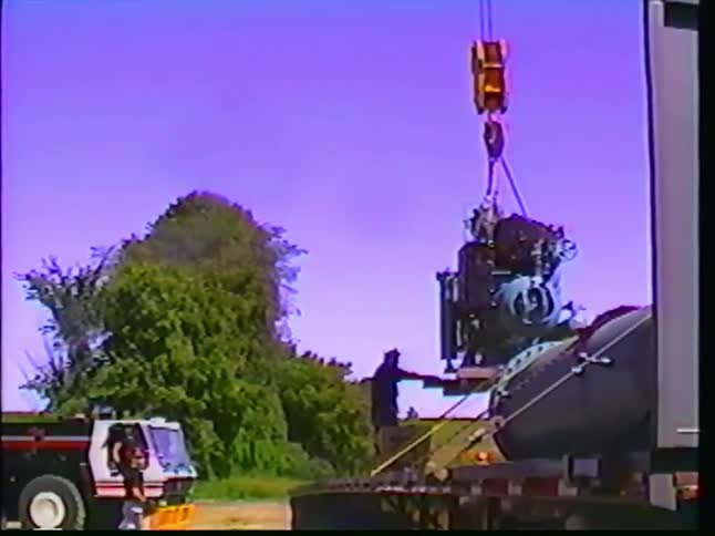 Retro VHS footage shows workers using a crane to carefully lift a large piece of industrial machinery from a flatbed truck
