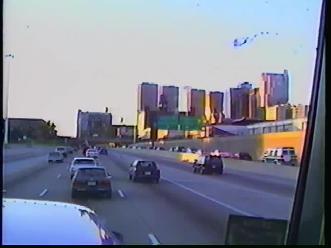 Vintage point-of-view footage captures a drive on a highway with traffic approaching a city skyline