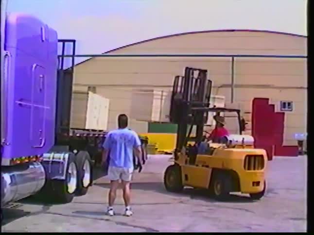 Vintage footage shows a yellow forklift loading pallets onto a distinctive purple semi-truck at a warehouse