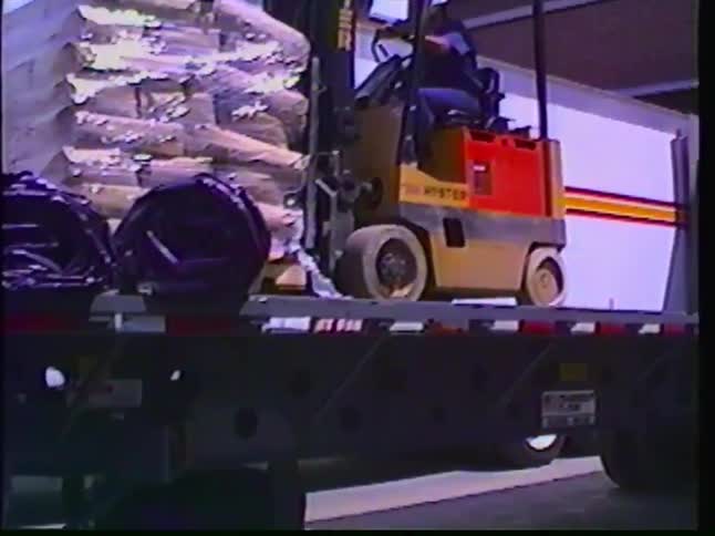 A forklift operator moves a pallet of shrink-wrapped bags off a flatbed truck and into a warehouse loading bay