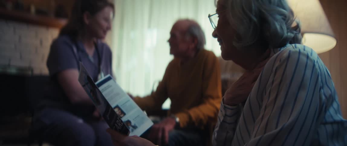 A home healthcare nurse explains a comprehensive care plan to an elderly couple using a brochure in their living room