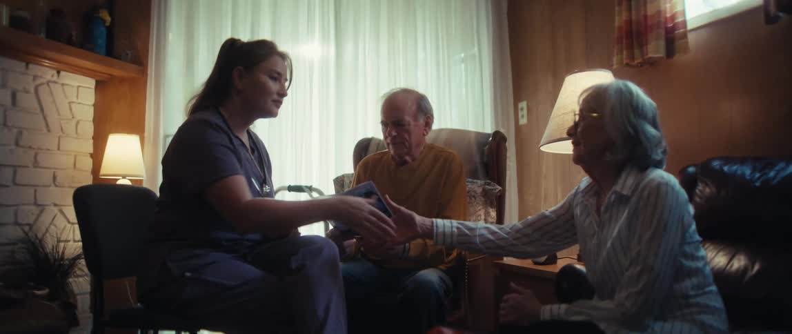 A friendly caregiver explains healthcare options from a brochure to an elderly couple during a home visit, ending with a warm handshake