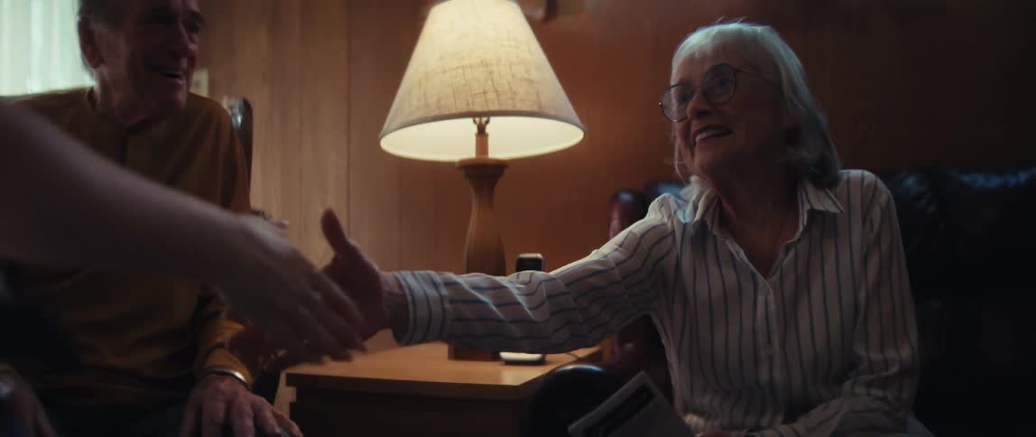 An elderly couple smiles warmly as they greet and shake hands with their female in-home caregiver in their cozy living room