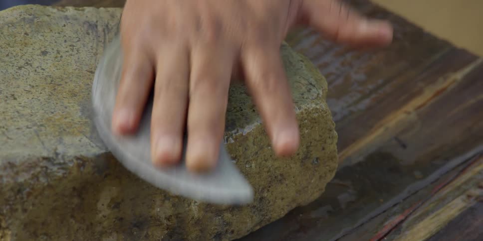 A craftsman hones the edge of a curved blade against a rough whetstone in a detailed close-up shot