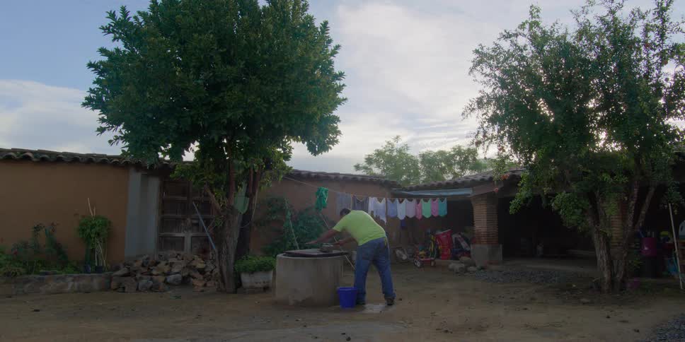 A man in a green shirt washes items by hand at a well in a rustic rural courtyard