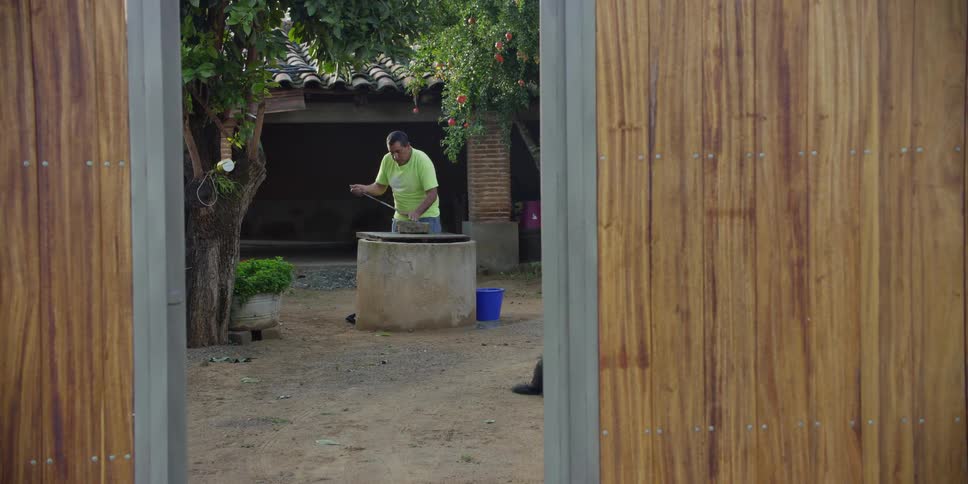A man in a rural courtyard sharpens a long machete on a stone over a well, framed by a wooden gate