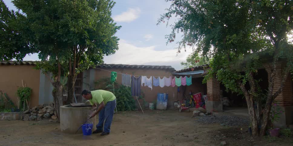 In a rural village yard, a man washes his clothes by hand at a well and hangs them to dry on a clothesline