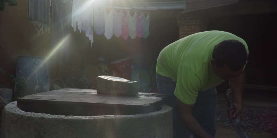 A man in a green shirt carefully cleans his machete by a stone well as the sun shines brightly in a rustic courtyard
