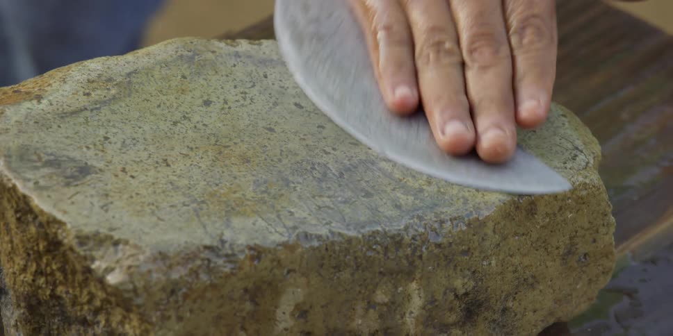 A craftsman meticulously sharpens a large curved blade on a wet whetstone in a close-up shot