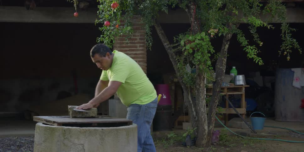 A Hispanic man in a bright green shirt carefully sharpens a large machete blade on a stone in his backyard