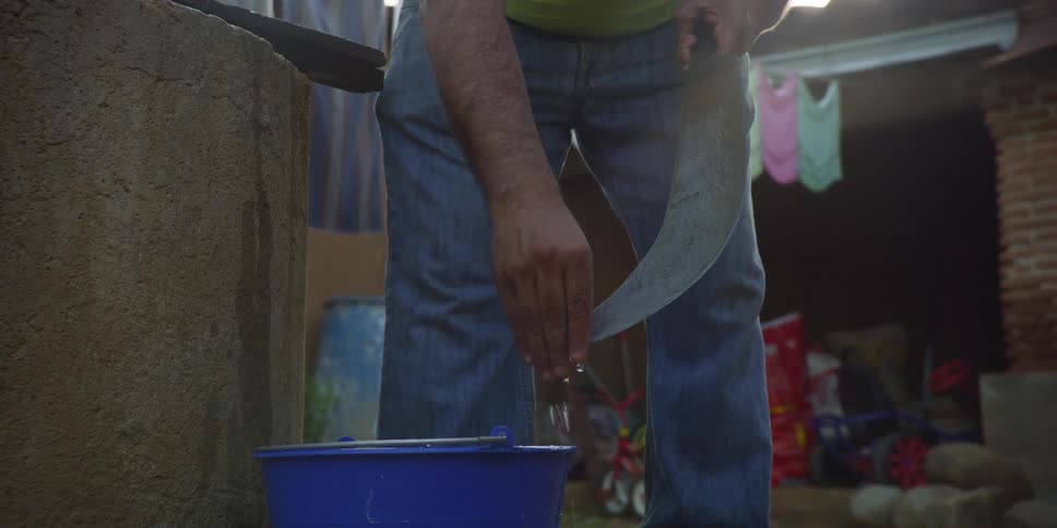 A man in jeans meticulously cleans a large, curved blade with water from a bucket in slow motion