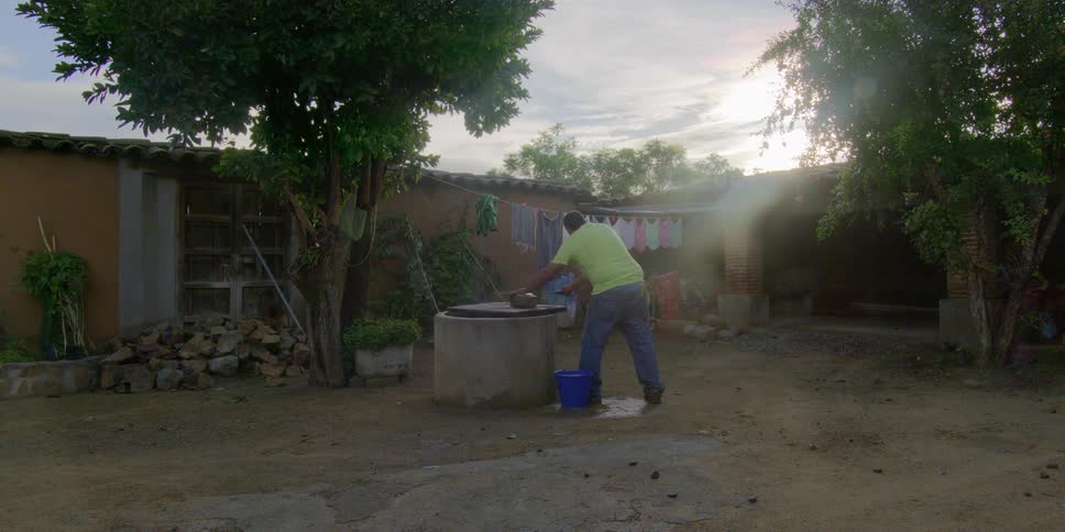 A man in a lime green shirt does chores at a concrete water well in a rustic backyard as the sun sets