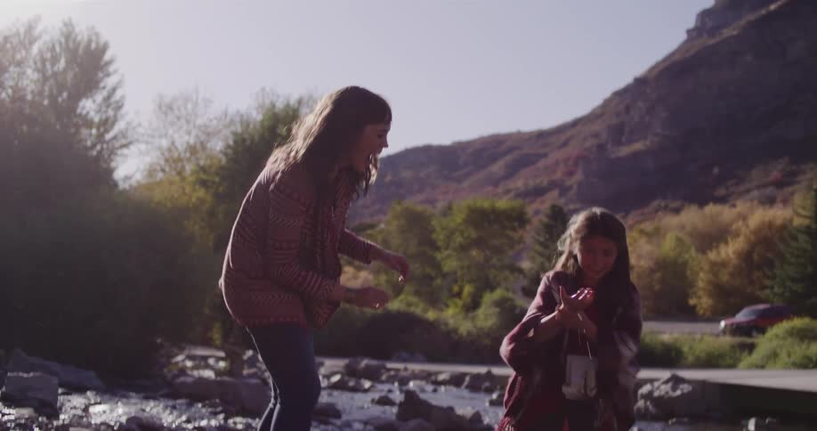 Playful mother and daughter laugh and splash each other with water in a sunlit mountain creek in slow motion