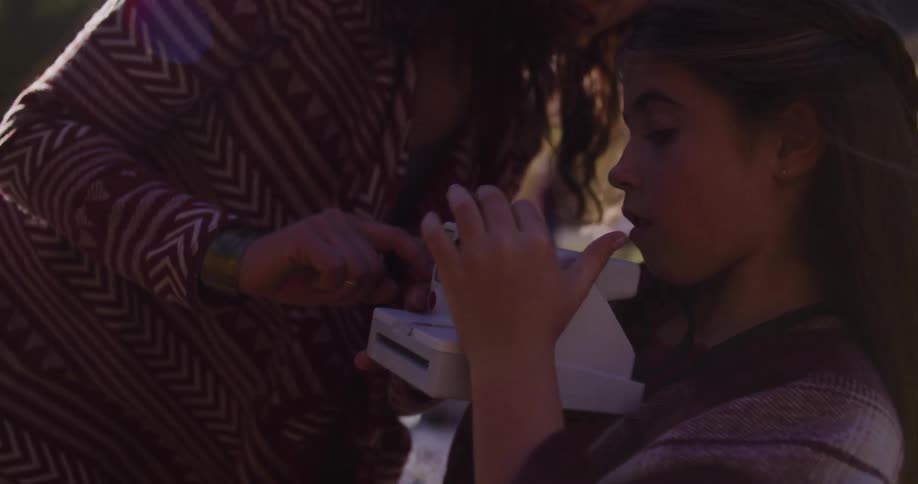 A mother teaches her daughter how to use a vintage instant camera in a sun-drenched outdoor setting