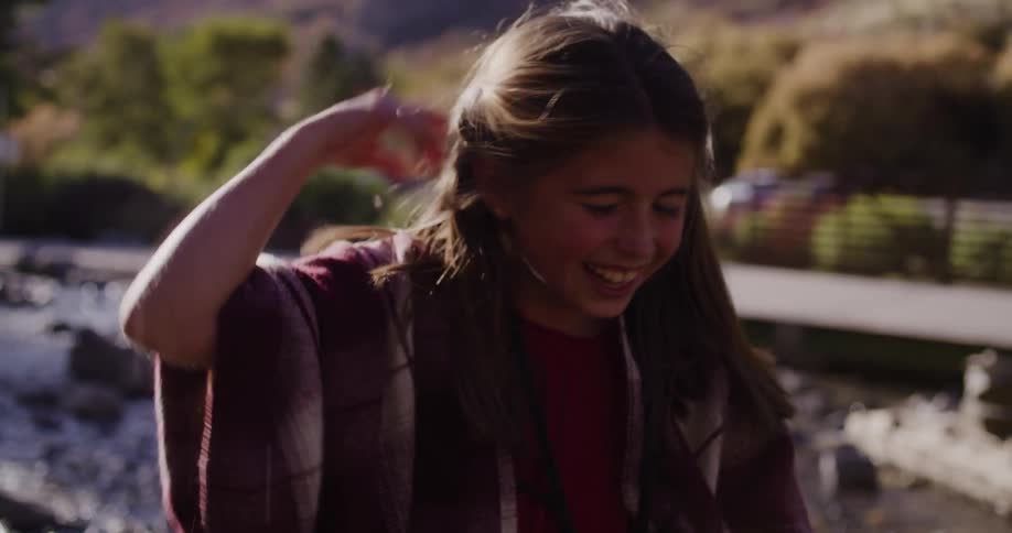 A joyful young girl wrapped in a plaid poncho laughs while splashing water in a sunlit river in slow motion
