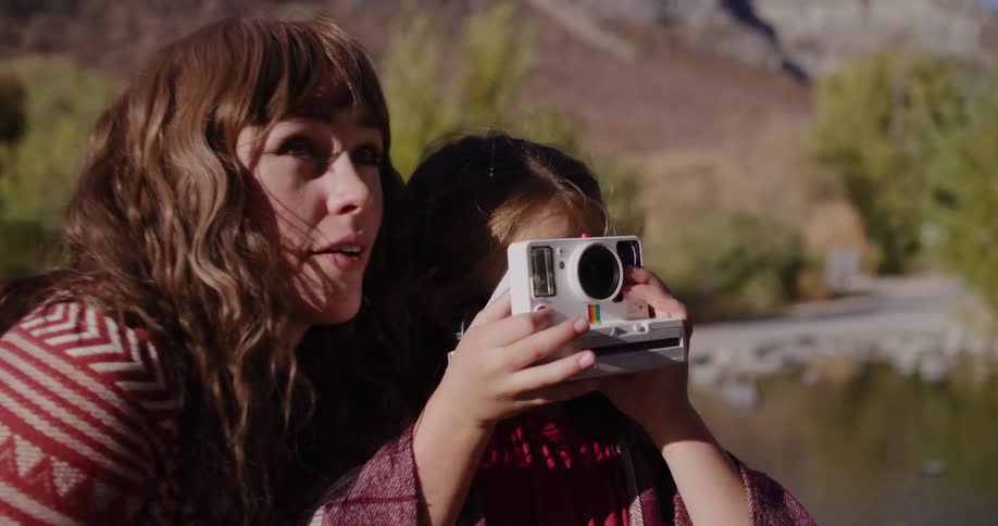 A mother teaches her daughter how to use a vintage instant camera, pointing out subjects as they capture memories in a sunlit canyon