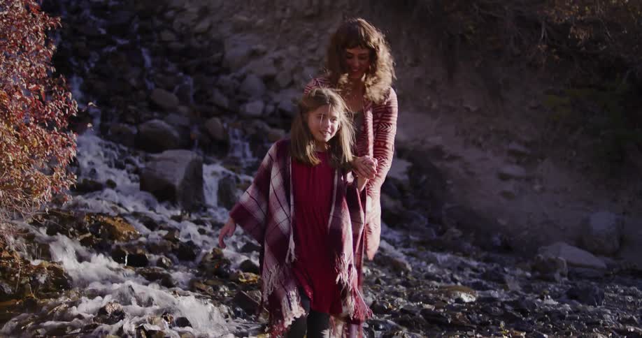 A caring mother helps her young daughter carefully walk across a shallow, rocky stream on a sunny day
