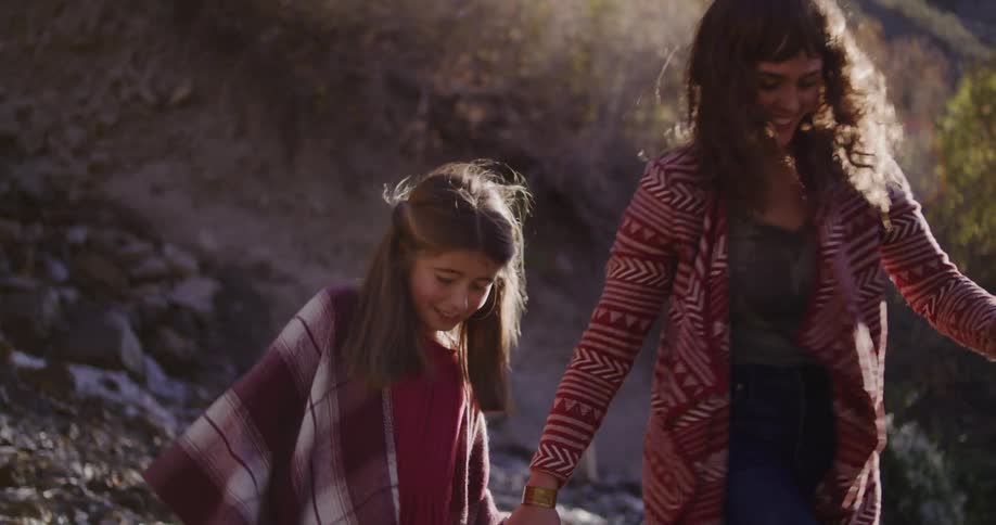 A mother and daughter hold hands and laugh while carefully walking along a rocky riverbed in slow motion