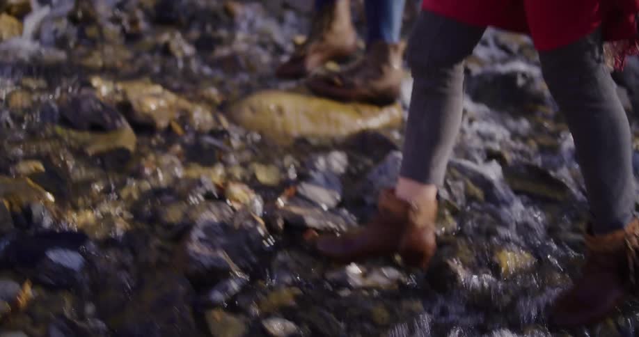A close-up slow-motion view shows a person's feet in brown leather boots carefully stepping across a rocky, flowing stream