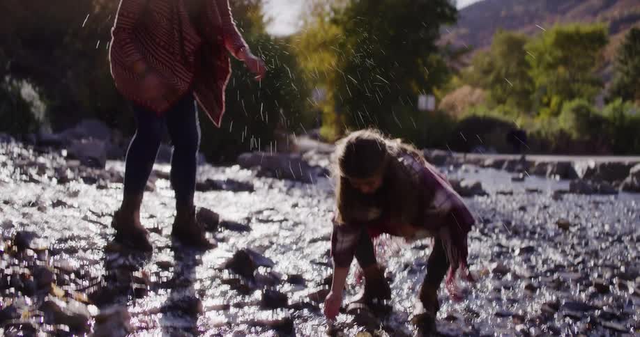 Mother and daughter playfully splash each other with water while standing in a shallow, sunlit river in slow motion