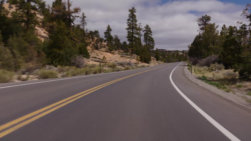 A point-of-view shot driving along a winding two-lane highway through a scenic high-desert landscape with pine trees and rocky hills