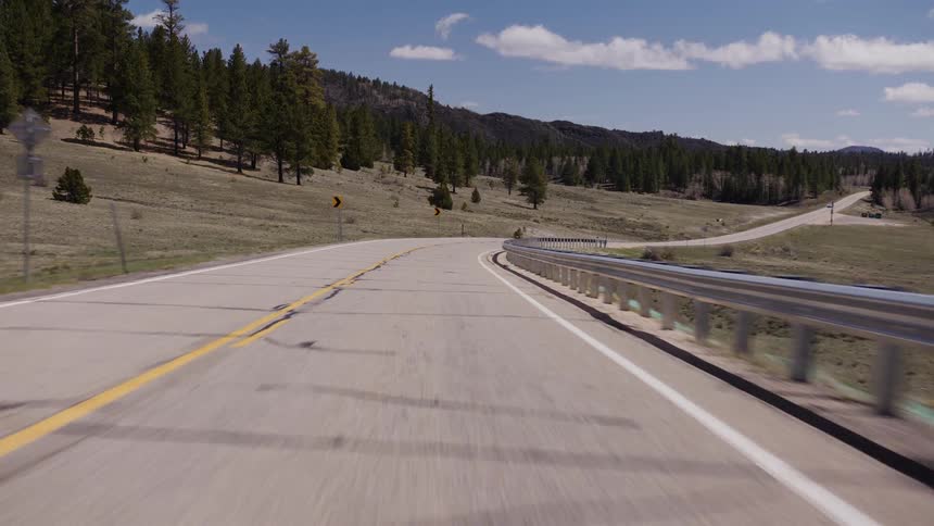 A point-of-view shot follows a winding rural highway through a scenic mountain landscape with pine trees on a bright sunny day