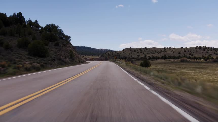 A point-of-view shot driving along a winding two-lane highway through a vast, hilly landscape under a clear blue sky